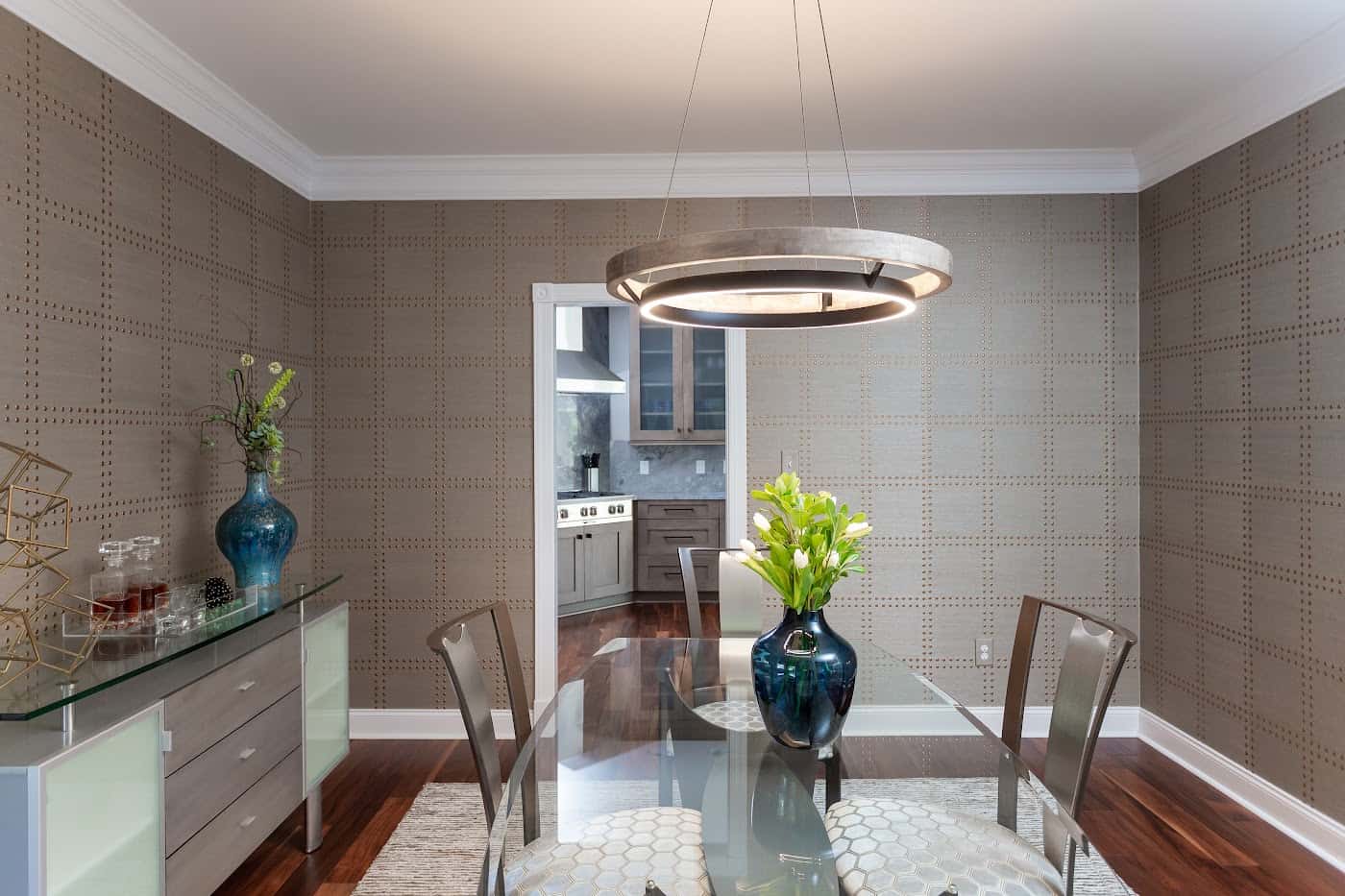 Dining room with glass table, ring chandelier, textured wallcovering and teal vase